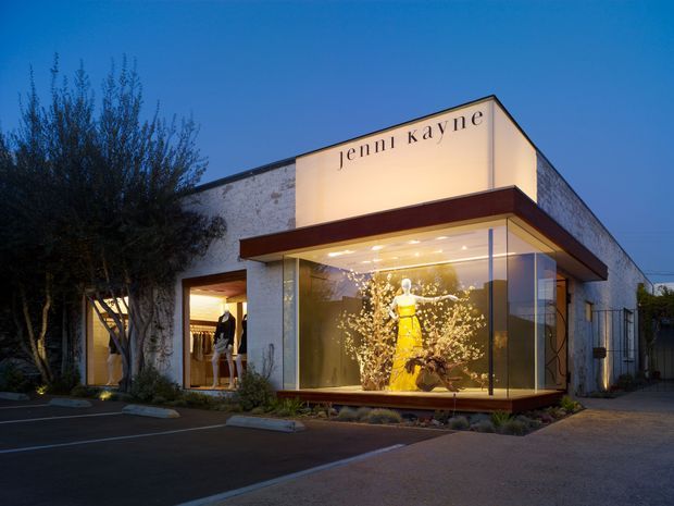 A dusk exterior photograph of a contemporary commercial retail or showroom building. The low, rectilinear structure has a pale textured masonry facade, a projecting dark reddish-brown canopy, extensive floor-to-ceiling glass shopfront glazing, and illuminated fashion window displays. The building signage reads “jennikayne,” with landscaped planting and a small parking area in front.