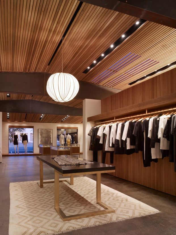 A warm, refined fashion retail interior with extensive timber slatted wall and ceiling linings, dark exposed structural beams, recessed linear lighting, and a large spherical pendant light. Clothing is displayed on open wall-mounted rails, with a central dark display table on a patterned neutral rug. The space extends toward a branded display area at the rear.