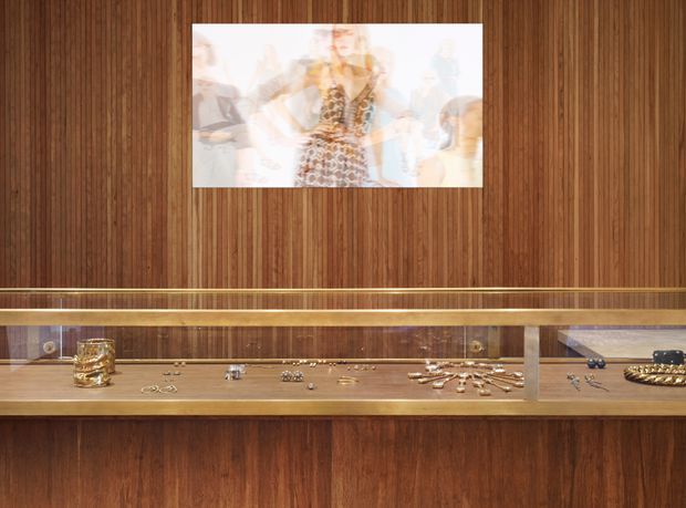 A refined jewellery display interior with a long, glass-fronted timber display case built into a richly grained dark wood wall. Jewellery is arranged across the illuminated brass-toned display shelf, with a fashion artwork mounted above. The composition is warm, orderly, and gallery-like, with strong horizontal lines and concealed display lighting.