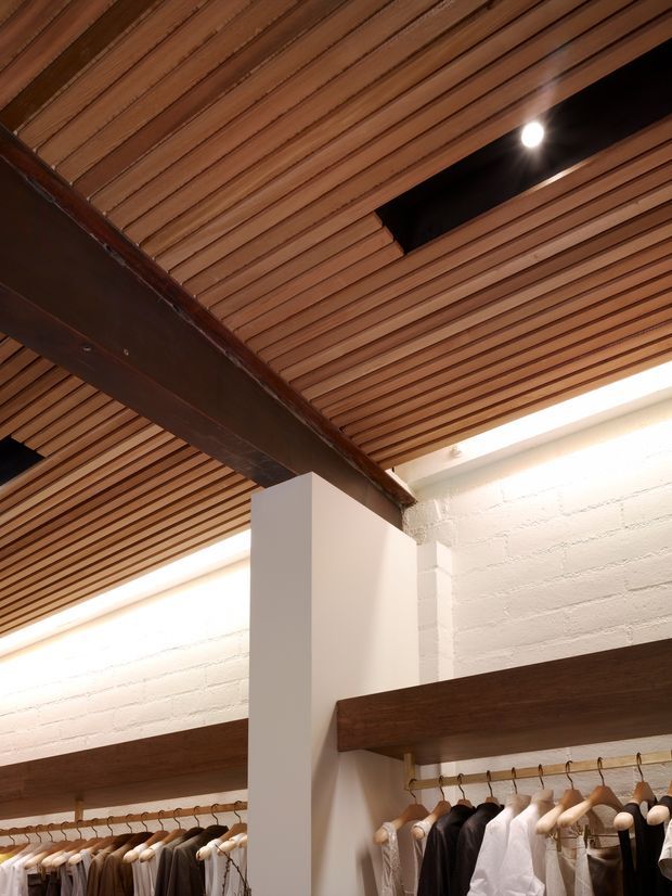 Interior architectural detail of a clothing retail space, featuring a warm slatted timber ceiling with recessed lighting and a dark exposed structural beam. White-painted brick walls, white structural columns, and dark timber clothing rails frame neatly displayed garments below.