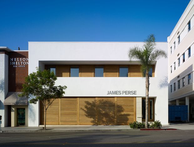 Street-facing commercial building with a clean white rendered facade, broad horizontal timber louvres screening the upper and lower glazing, and the James Perse name across the frontage. A neighboring Nelson Shelton real-estate office and a larger multi-storey building are visible at either side. Palm trees and a paved urban street reinforce the warm, contemporary retail setting.