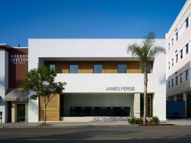 Street-facing view of a contemporary low-rise commercial building with a crisp white rendered facade, deep rectangular openings, timber-toned horizontal screening and doors, extensive ground-floor glazing, and the James Perse retail frontage. A taller modern building adjoins the right side, with palm trees and street elements in front.