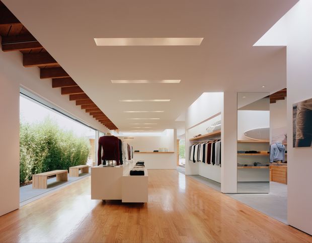 A bright, spacious contemporary clothing retail interior with polished timber flooring, extensive floor-to-ceiling glazing, white walls and ceiling, recessed linear lighting, exposed dark timber ceiling joists along one side, built-in clothing racks, shelving, display plinths and a large full-height mirror. A landscaped exterior is visible through the glazing.