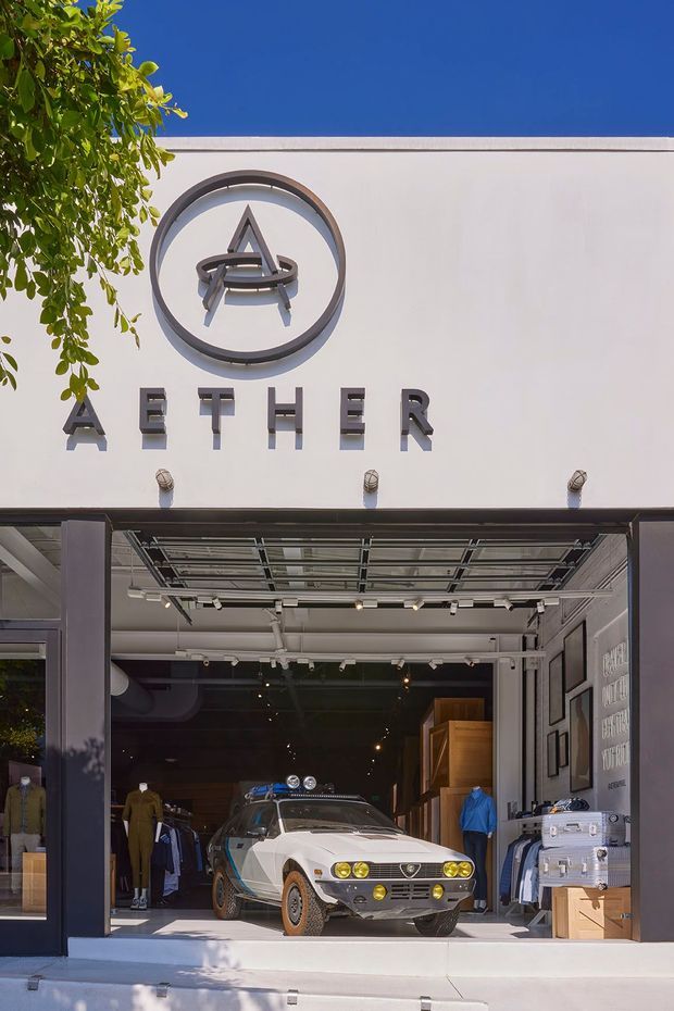 Street-facing storefront facade for Aether, with a large circular logo and raised lettering above a wide open glazed entrance. The contemporary white facade has dark metal framing, an exposed ceiling and track lighting inside, with a vintage sports car and clothing displays visible in the showroom.