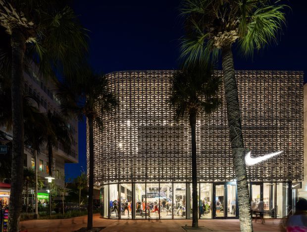 Night-time exterior photograph of a multi-storey urban retail building. The facade is wrapped in a distinctive illuminated geometric perforated screen, with a fully glazed ground-floor storefront and a large illuminated Nike swoosh sign. Palm trees and pedestrian paving frame the building in a tropical streetscape.