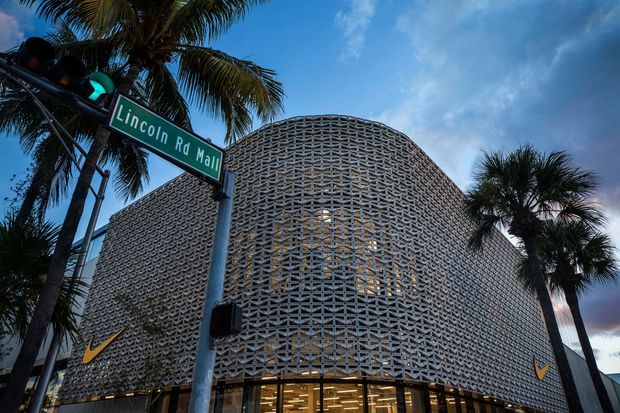 Exterior view of a contemporary commercial building on a palm-lined urban street, featuring a prominent curved facade wrapped in an intricate repeating breeze-block or perforated masonry screen. Large glazed retail-style openings occupy the ground level, with yellow swoosh logos visible on the facade. A Lincoln Road Mall street sign and tropical landscaping establish the streetscape context.