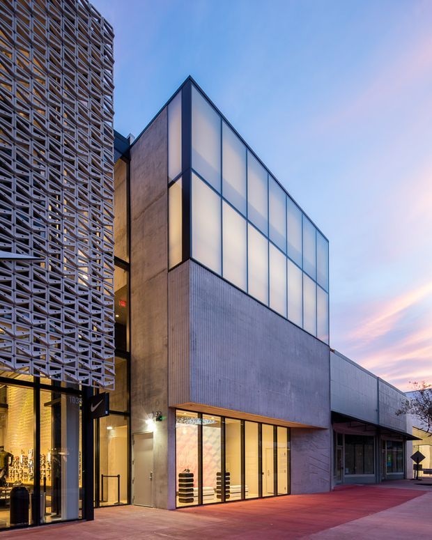 Exterior architectural photograph of a contemporary multi-storey commercial building at dusk. The facade combines a perforated geometric screen on the left, exposed concrete and vertically ribbed metal-like cladding, and a prominent upper volume enclosed in translucent grid-framed glazing. Ground-level retail or commercial glazing opens onto a broad red-toned paved forecourt, with warm interior lighting visible.