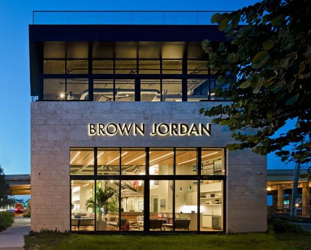A dusk exterior photograph of a contemporary two-storey commercial building, with a pale stone-clad facade, extensive black-framed glazing, illuminated interior spaces, and the prominent “BROWN JORDAN” sign. The upper level appears to be an open office or showroom, while the ground floor displays furniture and retail or commercial interiors. Landscaping and a covered structure are visible around the building.