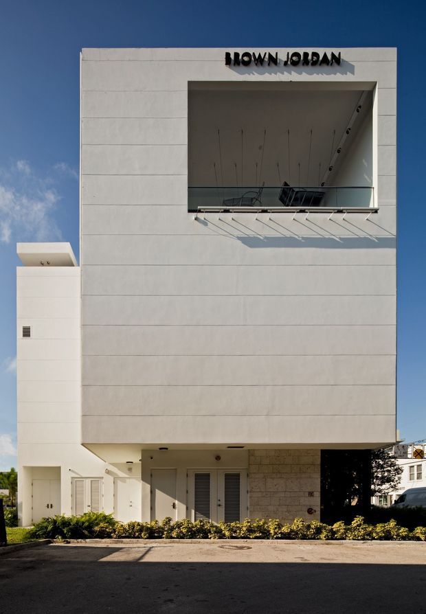 Front exterior view of a contemporary multi-storey commercial building with a clean white panelled facade, large recessed upper-level opening with glass balustrade, projecting sun-shading rods, and the “BROWN JORDAN” sign. The ground level includes recessed entrances, louvred doors, light stone cladding, landscaped planting, and a paved forecourt.
