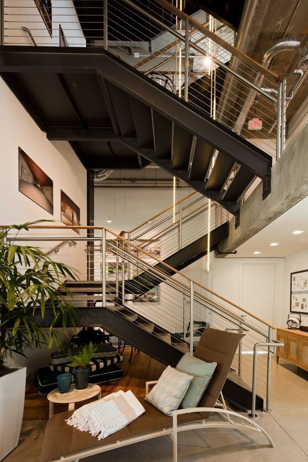 Interior view of a tall, industrial-style multi-level space centered on a dramatic black steel stair system with open risers and cable railings. Exposed ductwork, structural beams, concrete surfaces, timber handrails, and high ceilings create a warehouse-like character. A furnished lounge area with leather sling chairs, cushions, a small round table, indoor plants, and artwork occupies the foreground beneath the stairs.