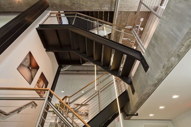 Looking upward through a tall interior stairwell, showing multiple dark steel stair flights and landings with slim metal balustrades. Exposed concrete walls and ceiling, visible services, linear lighting, and framed wall artwork create an industrial, multi-level circulation space.