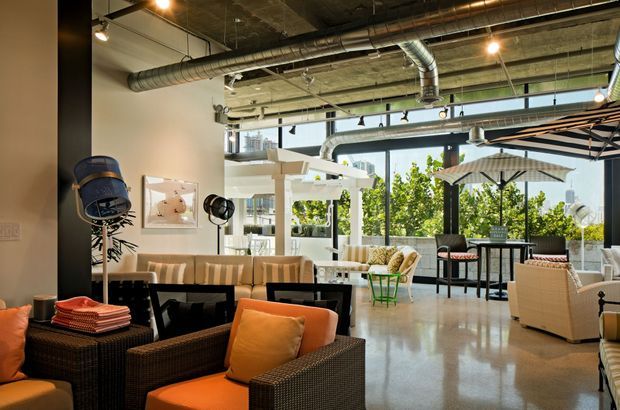 A bright, open-plan commercial lounge or hospitality interior with polished concrete flooring, exposed concrete ceilings, ductwork and track lighting. The space contains woven lounge chairs and sofas with orange cushions, café-style tables, bar-height seating, umbrellas, and extensive black-framed glazing opening toward a terrace or outdoor area. The overall aesthetic is industrial with warm contemporary furnishings and abundant natural light.