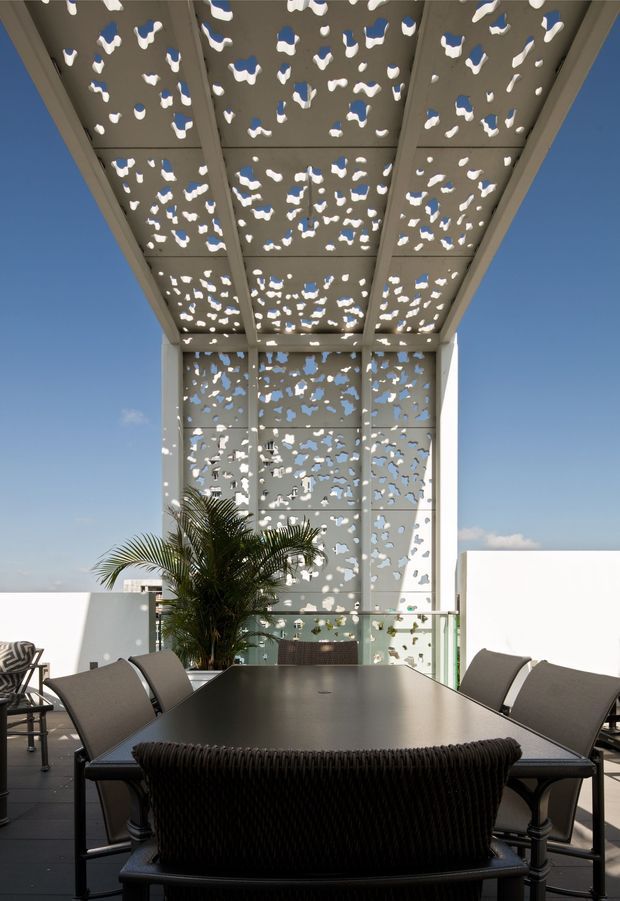 Outdoor rooftop or elevated terrace dining area with a large dark rectangular table and contemporary outdoor chairs, enclosed by a glass balustrade. A tall perforated metal screen and matching overhead canopy create a patterned architectural enclosure, with strong sunlight casting irregular shadows and openings against a clear blue sky. A palm adds greenery, while white parapet walls and dark decking complete the space.