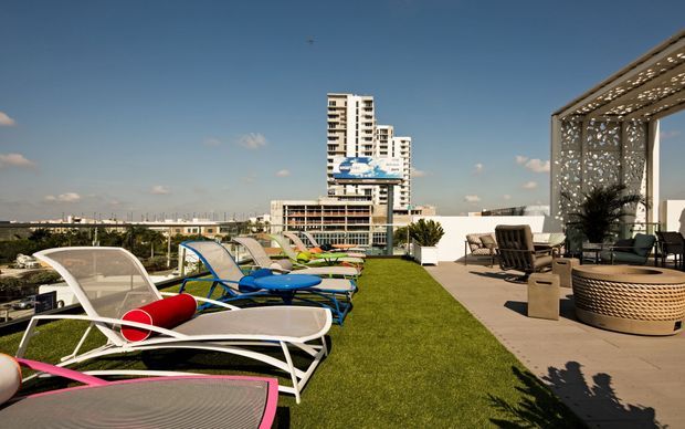 A spacious rooftop terrace with artificial lawn, colourful modern pool loungers, and a covered outdoor lounge area furnished with low seating, a round woven table, and potted plants. The terrace overlooks a dense urban skyline, including several high-rise buildings, under a clear blue sky. The setting has a relaxed resort-like atmosphere with strong mid-century-inspired outdoor furniture and contemporary rooftop architecture.