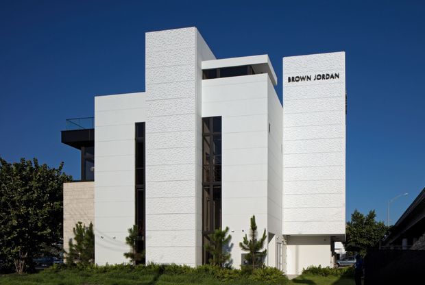 Exterior view of a modern multi-storey commercial or professional building with a stark white geometric facade. The composition features tall textured white vertical wall planes, smooth white panelled surfaces, narrow vertical glazing, a projecting dark balcony or terrace, and a recessed central entry zone. The building is set against a clear blue sky with landscaping at ground level, and the words “BROWN JORDAN” are displayed prominently on the right facade.