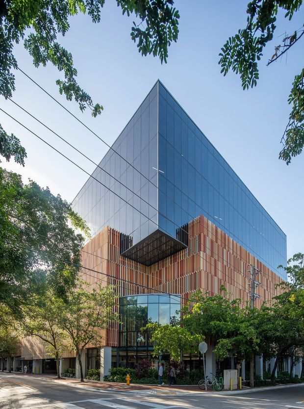 Street-level exterior view of a large contemporary multi-storey building with a sharply angled, glass-clad upper volume above a warm timber-toned vertical screen facade. The building includes projecting geometric features, a glazed entrance or lower-level frontage, and mature street trees along the urban streetscape.