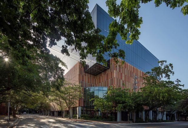 Exterior architectural photograph of a multi-storey contemporary building set along a tree-lined urban street. The facade combines extensive blue-grey glazed curtain walling with a lower section of vertical timber-toned fins and recessed balconies or canopies. Mature trees partially screen the building, while a glazed ground-level entrance and shaded street frontage are visible.