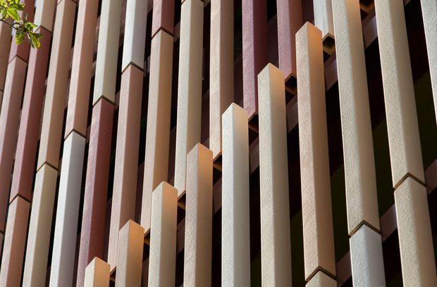 Close-up architectural detail of a facade composed of repeated tall, narrow vertical fins or pilasters in alternating cream, pale pink, burgundy, and muted orange tones. The textured projecting elements create strong rhythm, depth, and geometric shadow lines, with a small amount of foliage visible at the upper left.