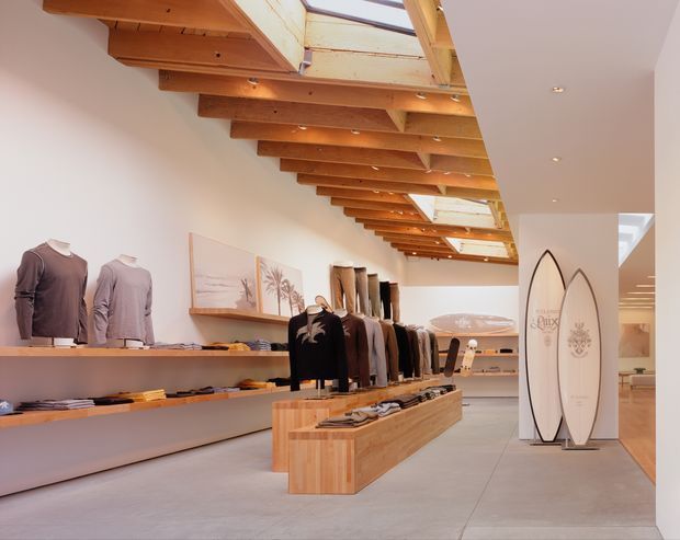 Bright contemporary retail showroom with clothing displayed on wall-mounted shelves and central timber display joinery. The space features a long skylight with exposed warm timber beams and rafters, recessed ceiling lighting, polished pale concrete flooring, neutral plaster walls, framed artwork, and surfboards displayed near the rear circulation area.