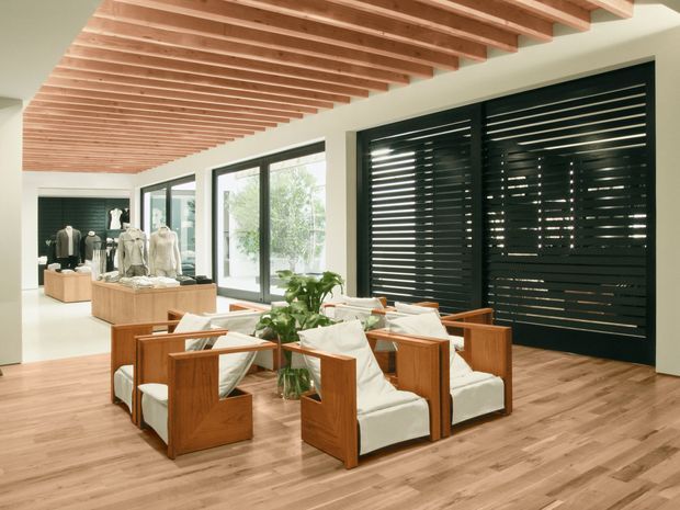 A bright, contemporary retail showroom or boutique interior with a central arrangement of low-profile timber-framed lounge chairs upholstered in white. The space features warm timber flooring, a dramatic exposed timber slat ceiling, large windows, and a dark louvred window or door screen along one wall. Clothing displays are visible in the background, confirming a commercial showroom setting. Indoor plants and light-coloured walls add a clean, natural feel.