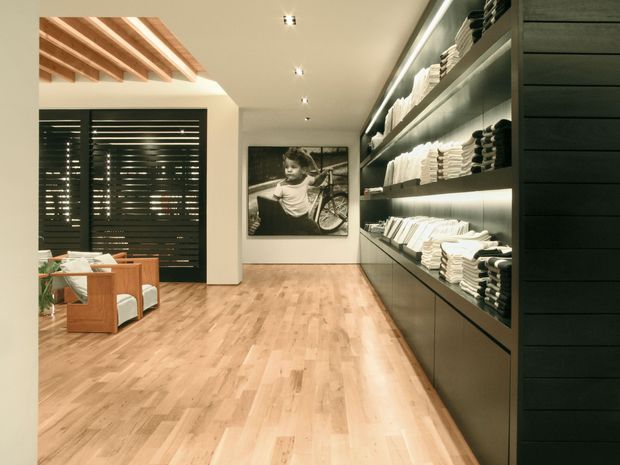 A refined retail or showroom interior with a long wall of dark built-in shelving displaying neatly folded textiles, illuminated by integrated strip lighting. The space has a warm timber floor, white walls, dark horizontal window blinds, exposed timber ceiling beams over a seating area, and a large black-and-white photographic artwork.