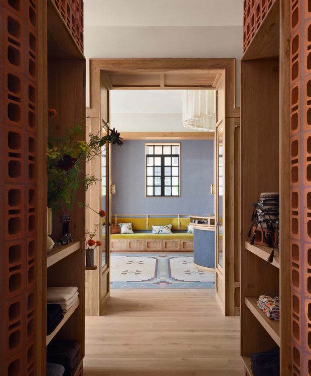 View through a warm timber-lined passage framed by open shelving and distinctive terracotta breeze-block screens, looking into a colourful living room. The room beyond has a blue-grey wall, mustard built-in window seat, patterned rug, black-framed window, and a sculptural hanging light. The composition highlights layered thresholds, integrated joinery, and textured masonry screens.