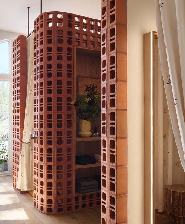 Interior view of a warm, light-filled room featuring a prominent freestanding open shelving and partition structure built from exposed terracotta hollow bricks. The brickwork forms curved screens and vertical columns around recessed timber shelving containing plants and folded textiles. Pale walls, timber flooring, sheer patterned curtains, and a partial timber-framed opening create a relaxed, earthy atmosphere.