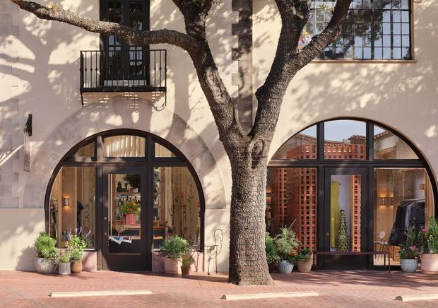 Street-facing facade of a refined low-rise commercial or mixed-use building, with pale stucco walls, large arched black-framed glazed openings, a small wrought-metal balcony, and a mature tree integrated into the foreground. The arched storefronts reveal a warmly styled retail or hospitality interior, while brick paving and potted plants create an inviting urban frontage.