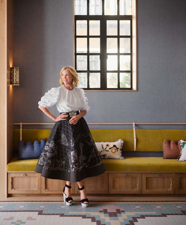 A stylish lounge or reception area with a long built-in mustard-green upholstered bench over timber cabinetry, set against dark textured walls. A large black-framed multi-pane window provides natural light, while a decorative wall sconce and patterned pillows add visual interest. The floor features a geometric multicoloured tile pattern. A woman stands in the foreground, but the architectural focus is the built-in seating and interior finishes.