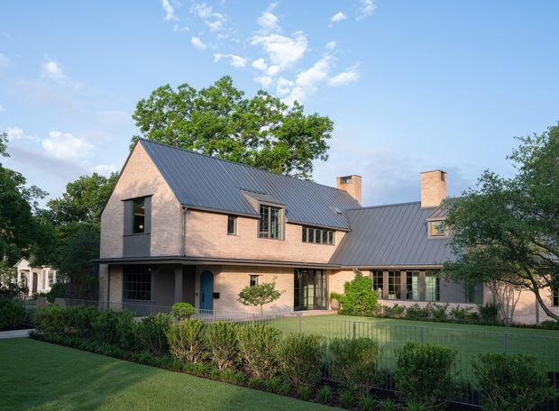 Exterior view of a substantial two-storey brick house with steep gabled and intersecting metal roofs, tall brick chimneys, dark-framed windows, and a covered entry terrace. The home is set within a landscaped suburban garden with hedges and lawn under a bright blue sky.