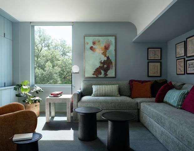 A refined living room with a large grey sectional sofa, two black cylindrical coffee tables, and a pair of warm brown lounge chairs. The room features pale blue built-in cabinetry, a matching curved-edge blue-grey feature wall, a large window overlooking trees, framed artwork, layered cushions in burgundy, rust, and teal, and a potted plant. Natural daylight creates a calm yet richly coloured contemporary interior.