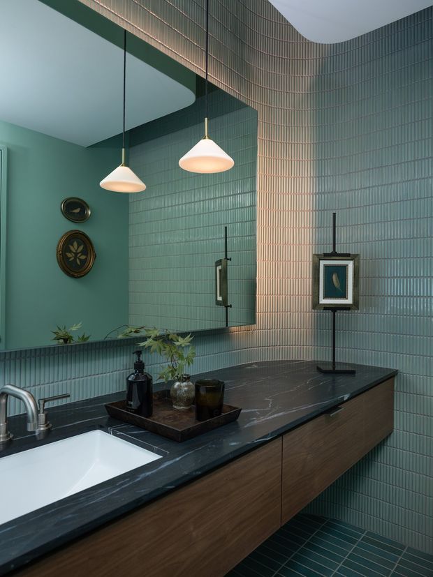 A refined bathroom vanity with a long dark stone benchtop, integrated white basin, and warm timber floating cabinetry. The walls and floor are clad in narrow glossy green tiles, wrapping around a curved corner and reflected in a large mirror. Two sculptural pendant lights hang above the vanity, while framed artwork and small decorative objects add character. The palette is deep green, charcoal, timber brown, and warm white, creating a moody yet elegant interior.