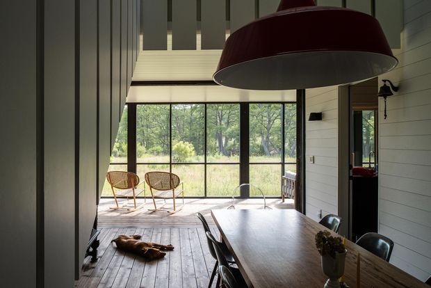 A dark, refined dining area opens onto a bright glazed sunroom or covered outdoor deck. The room features a long timber dining table with black chairs, a large sculptural pendant light, timber plank flooring, and white horizontal wall panelling. Floor-to-ceiling glazing frames a wooded landscape, while woven lounge chairs sit in the adjoining sunlit space.