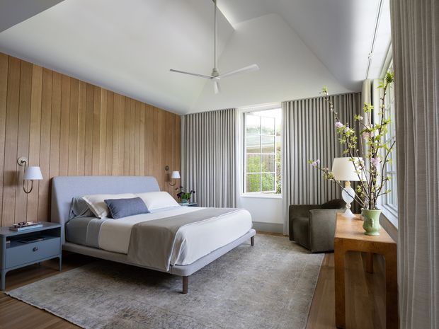A bright, refined bedroom with a low upholstered bed, layered white and pale blue bedding, and a large neutral rug. The room features warm vertical timber wall panelling on one side, grey vertically ribbed wall treatment around the window, a high angled white ceiling with a minimalist ceiling fan, and full-height curtains. A small blue bedside table, wall-mounted lamps, an armchair, and a timber console with a vase and branches complete the calm contemporary interior.