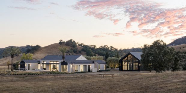 A wide dusk exterior view of a contemporary rural residence set among rolling dry hills. The single-storey home has a long, low horizontal composition, pale rendered walls, dark-framed glazing, multiple pitched roof forms, and a prominent glazed gable-ended outbuilding or pavilion to the right. Sparse trees and palms surround the house, with an open dry field in the foreground and hills behind.