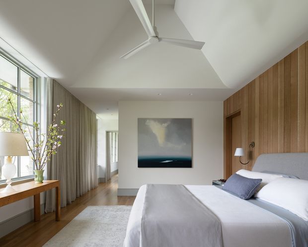 Bright contemporary bedroom with a pitched, angled ceiling and white ceiling fan. The room features full-height glazing with soft curtains, warm timber flooring, a vertical timber-panelled feature wall with integrated bedside lighting, a grey upholstered bed, pale bedding, and a light rug. A large abstract artwork and simple timber console complete the calm, refined interior.