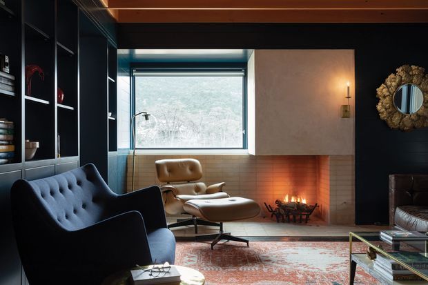 A moody, refined living room with a dark built-in shelving wall, deep blue tufted lounge chair, mid-century leather lounge chair and ottoman, patterned rug, glass coffee table, and a tiled fireplace with an active fire. A large window brings in a view of trees, while warm timber ceiling boards and dark walls create a dramatic, intimate atmosphere.