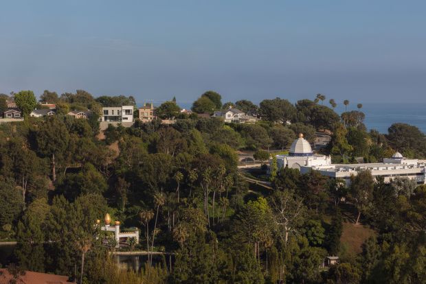 Elevated exterior view across a heavily wooded coastal hillside, with scattered residential buildings in the midground and a distinctive white institutional building with domed roof in the lower right. The ocean is visible beyond the treeline under a clear blue sky.