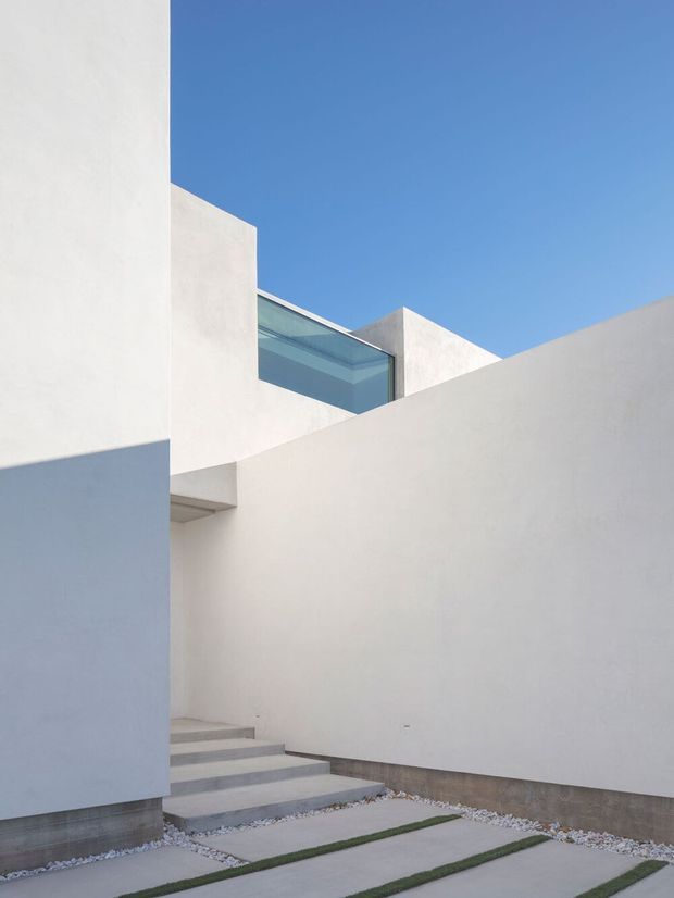 Minimalist modern courtyard with crisp white rendered walls, stepped concrete paving separated by narrow strips of grass and white gravel, and a glazed opening beneath angular geometric roof and wall forms. The composition emphasizes strong rectilinear volumes, shadow, and a clear blue sky.