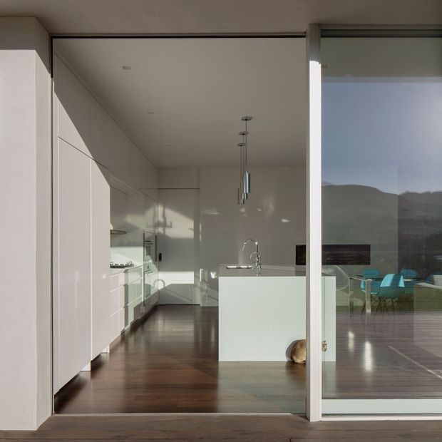 A minimalist contemporary kitchen viewed through a large glazed sliding opening. The room has white full-height cabinetry, a white island with integrated sink and curved faucet, stainless-steel pendant lights, dark timber flooring, and expansive glazing opening onto a timber deck with outdoor seating and mountainous scenery beyond.