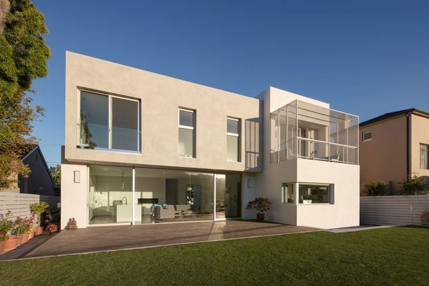 A contemporary two-storey residential home with a clean rectilinear form, pale rendered walls, extensive glazing, and a projecting upper-level balcony or enclosed terrace. The house opens onto a timber deck and manicured lawn, with neighbouring homes and mature planting visible around the site. The composition is bright and refined, featuring strong horizontal and vertical geometry.
