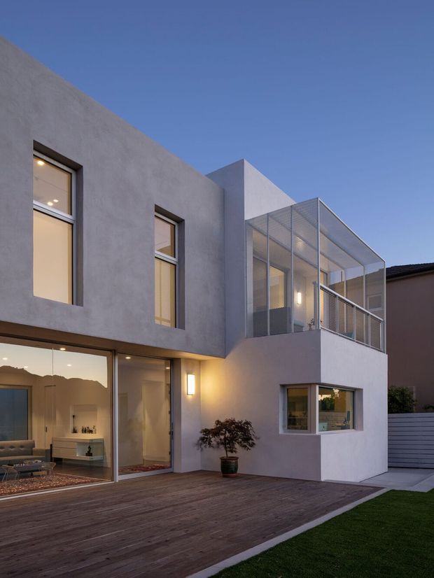 Twilight exterior view of a contemporary multi-level residence with smooth pale rendered walls, large glazed openings, a timber deck, and a prominent enclosed upper balcony with fine metal mesh screening. Warm interior and exterior lighting creates an inviting atmosphere, with a small lawn and side walkway visible.
