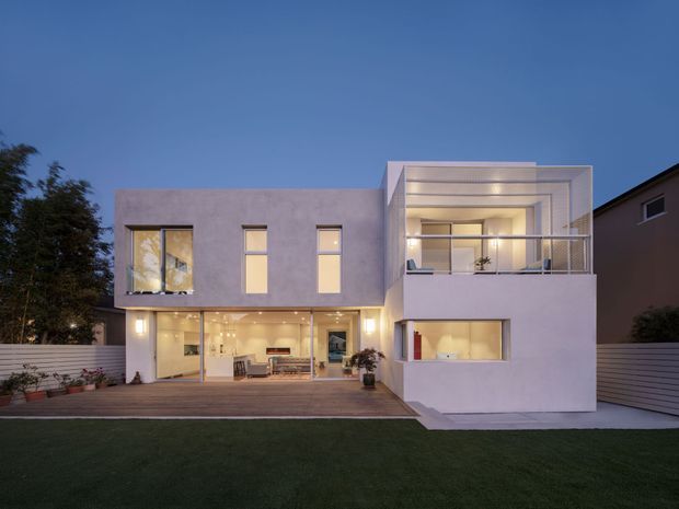 Twilight rear exterior view of a contemporary two-storey residence with a box-like rendered facade, flat rooflines, large glazed openings, a recessed upper balcony with metal balustrade, and a broad timber deck opening onto a lawn. Warm interior lighting is visible through the ground-floor glazing.