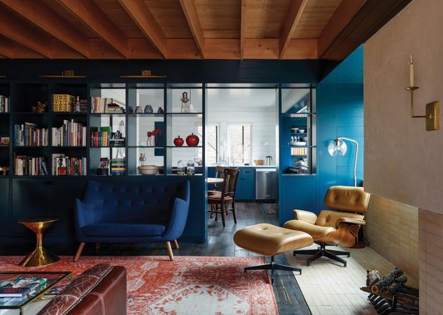 A richly layered open-plan living room with a dark navy built-in bookcase and room divider, a deep blue tufted sofa, and vintage leather lounge chairs with an ottoman. A compact dining area and blue kitchen cabinetry are visible beyond the glazed shelving. The room features a warm exposed timber-beam ceiling, pale brick and plaster walls, patterned Persian-style rugs, timber flooring, brass lighting and side tables, and a strongly curated mid-century-inspired palette.
