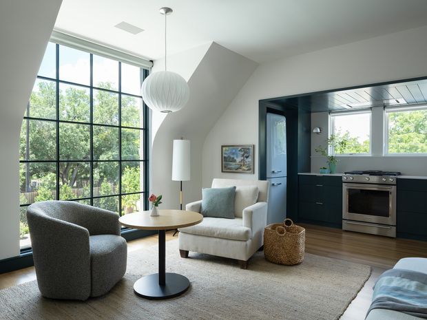 Bright, contemporary open-plan living room with a compact integrated kitchen. The room features expansive black-framed floor-to-ceiling glazing, a sculptural sloped ceiling, neutral upholstered seating, a round timber-topped pedestal table, a large textured rug, and a spherical pendant light. The adjacent kitchen has dark fixed cabinetry, a stainless-steel oven and rangehood, and a light tiled or paneled ceiling.