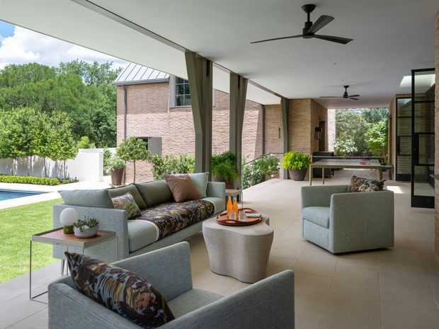 Covered outdoor living and entertaining area adjoining a brick-clad home, with a tiled floor, ceiling fans, large upholstered sofas and armchairs, a coffee table, dining table, and views toward a landscaped garden and swimming pool. The space is bright, open-sided, and suited to relaxed indoor-outdoor living.