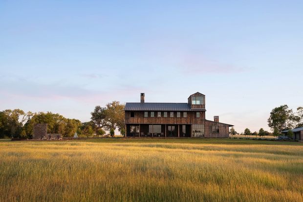 A large rural barn-style building set at the edge of an expansive golden field. The dark weathered timber and corrugated metal structure has a long rectangular form, multiple rows of windows, a small rooftop cupola, and attached side volumes. Mature trees and outbuildings sit in the background beneath a clear pastel evening sky.