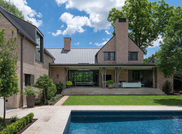 A refined brick residential exterior viewed from the backyard, featuring a pitched metal roof, prominent brick chimneys, large dark-framed glazing, and a covered outdoor living veranda. A paved terrace and rectangular swimming pool occupy the foreground, with manicured lawn and planting around the house. The image has a bright, clean contemporary-traditional character under a blue sky.