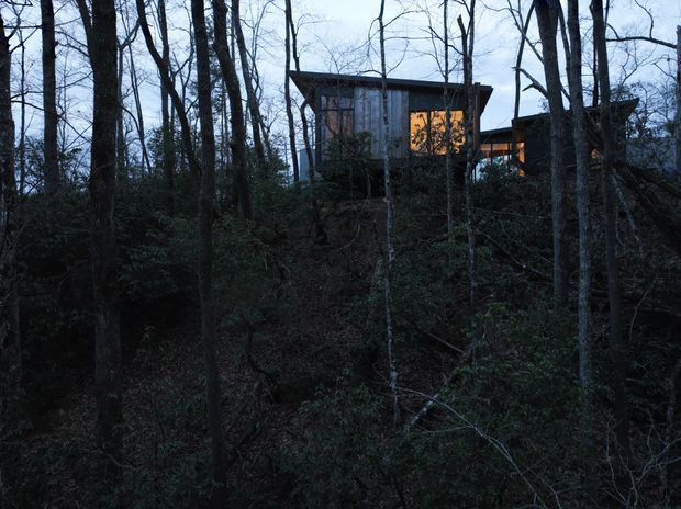 A dark, atmospheric exterior view of a modern cabin-like house partially concealed among dense leafless woodland. The elevated building has a simple angular flat or low-pitched roof, dark vertical cladding, large warm-lit windows, and a second connected or projecting volume visible to the right. Trees and undergrowth dominate the foreground.