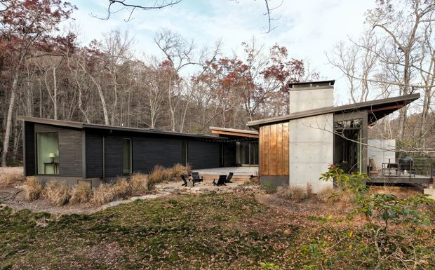 Single-storey modern residence set within a wooded landscape, composed of low horizontal dark-clad wings and a taller concrete volume. The house features broad overhanging roof planes, extensive glazing, warm timber cladding, and an outdoor terrace or patio area, with a restrained palette that integrates into the surrounding trees.